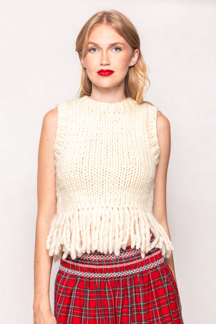 Wearing the Ivy City Co Gia Sweater in Cream, styled sleeveless with a chunky knit and thick yarn fringe hemline, a person is paired with a red plaid skirt and stands against a plain light background.