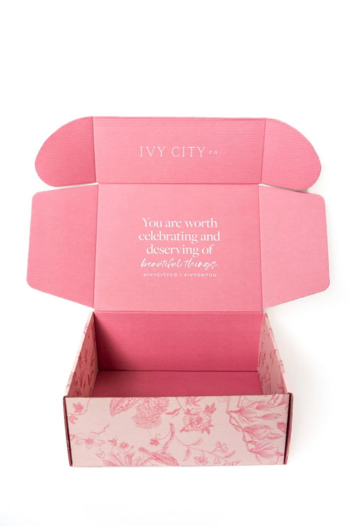 A beautiful Ivy City Co Gift Box is shown with the lid open, revealing a light pink interior and elegant floral patterns along the sides. Inside, a heartfelt message reads: “You are worth celebrating and deserving of beautiful things.” This personalized gift box is a thoughtful way to show you care.