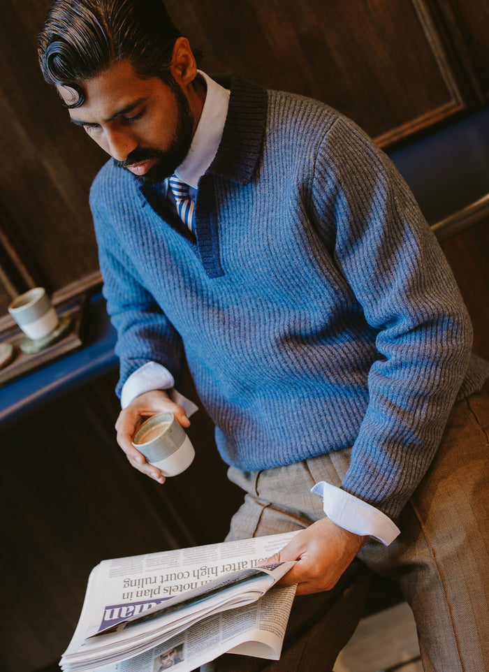 A model wears the Percival two-toned blue Picnic Polo, layered over a shirt and tie, reading a newspaper and drinking coffee.