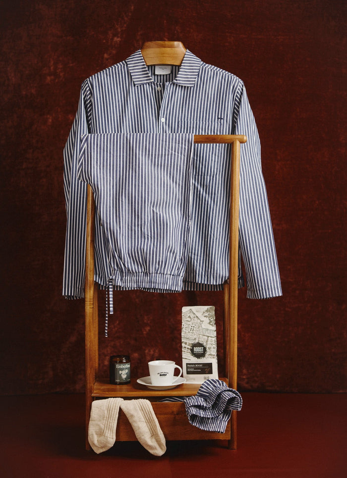 The Percival blue striped cotton pyjama set styled on a wooden valet stand with cosy morning essentials.