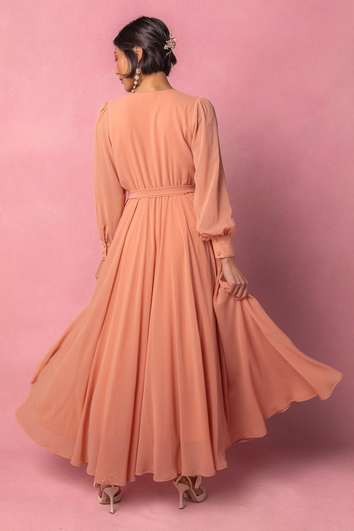 An Ivy City Co Andie Dress in Apricot Crush, featuring long sleeves and a flowy silhouette, is worn by a woman with dark hair styled in a low bun adorned with decorative hairpins. She stands against a pink background, facing away and holding the hem of her skirt, paired with beige heels.