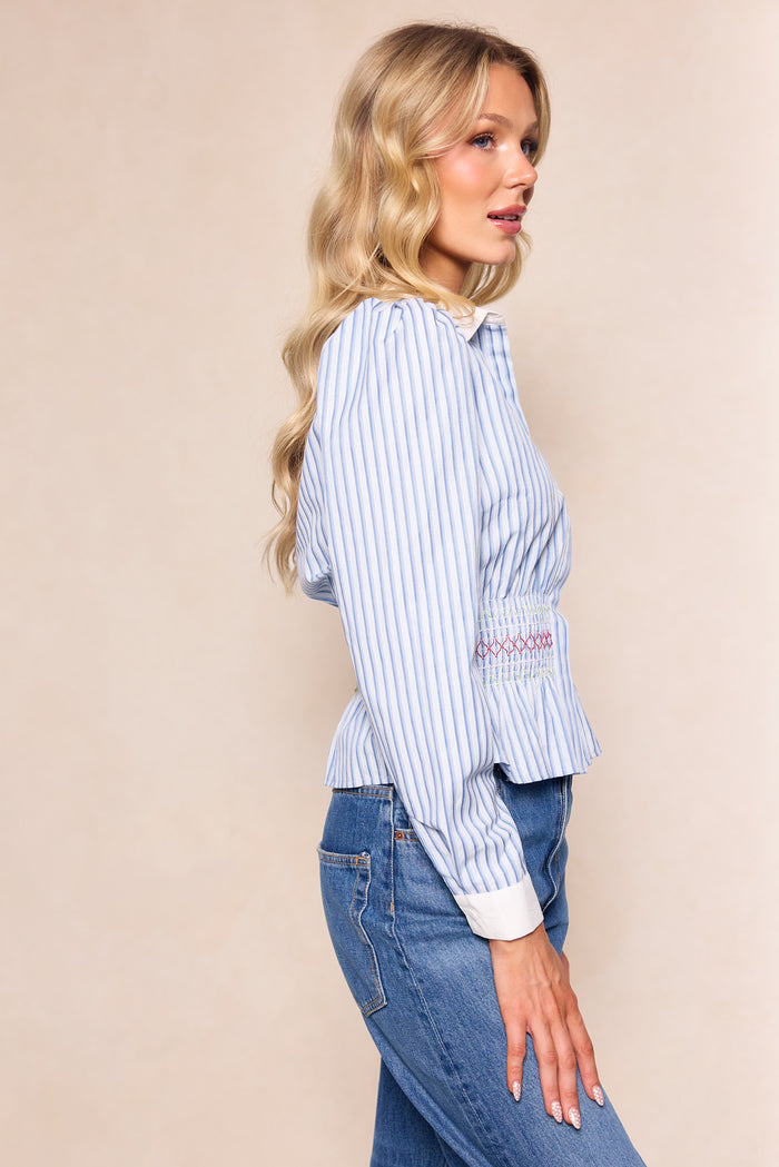 A woman with long wavy blonde hair wears Ivy City Co’s Bluebell Top in Blue & White Stripes, paired with blue jeans, standing sideways against a neutral beige background.