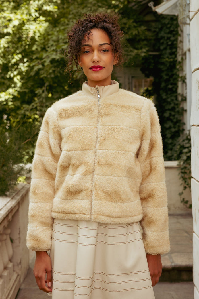A woman with curly hair is seen outdoors in front of greenery and a white building, wearing the Ivy City Co Brianna Jacket in Cream Fur paired with a cream skirt. She looks at the camera with a neutral expression. This stylish faux fur jacket is maternity friendly, delivering both elegance and comfort.