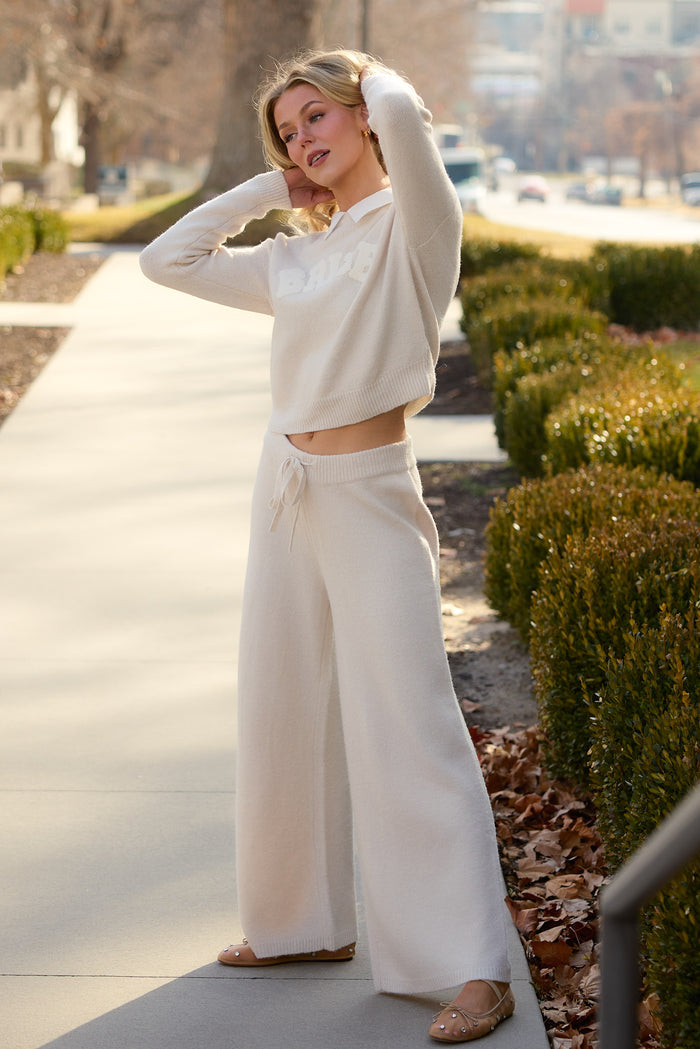 A woman stands outdoors on a sidewalk, wearing the Ivy City Co Bride Sweater in Oatmeal with wide-leg pants and flat shoes. Her hands are raised to her head, surrounded by trimmed bushes and dry leaves.