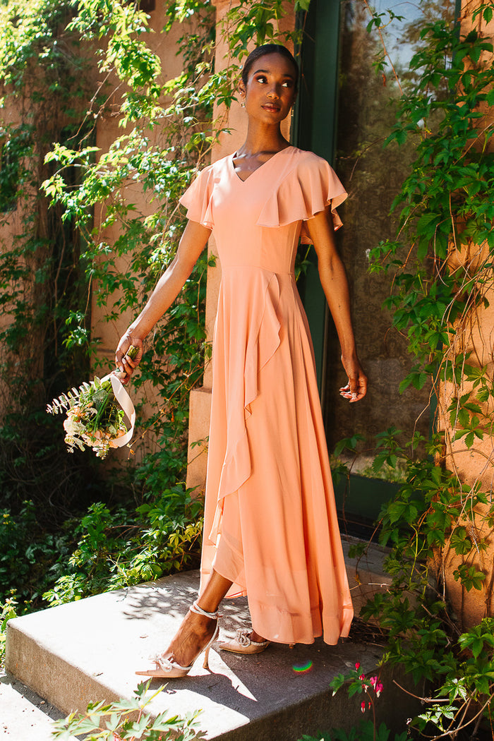 A woman in the Ivy City Co Callie Dress in Apricot Crush Chiffon with flutter sleeves stands outdoors by a vine-covered wall, holding a small bouquet and gazing upwards in warm sunlight.