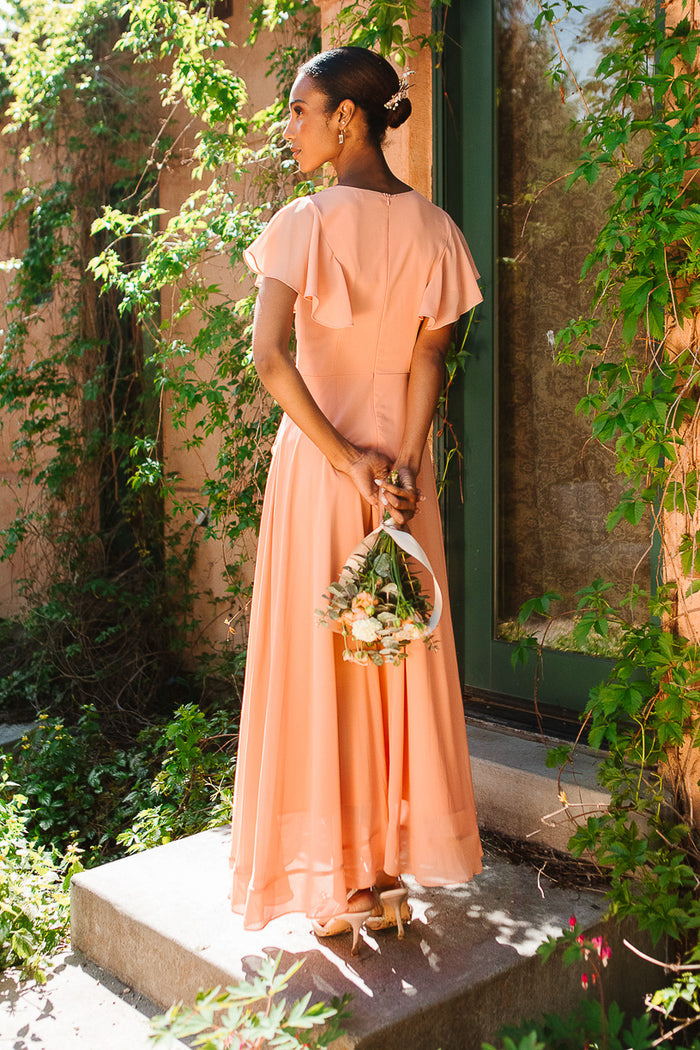 A woman wears the Ivy City Co Callie Dress in Apricot Crush Chiffon, standing on sunny concrete steps with greenery, holding a small bouquet behind her back and facing away from the camera.