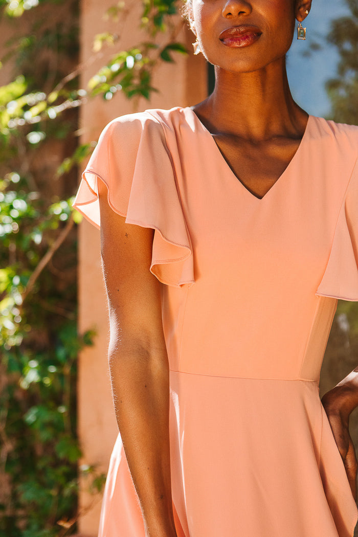 A woman stands outdoors, partially shaded by greenery, wearing the Ivy City Co Callie Dress in Apricot Crush Chiffon. Only her face and upper body are visible, glowing softly in the sunlight.