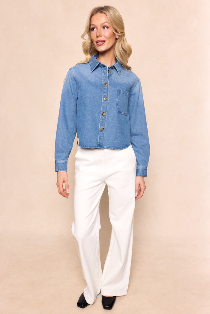 A woman with blonde wavy hair smiles softly against a beige backdrop, wearing Ivy City Co's Caroline Top in Light Wash Denim—a nursing-friendly style—paired with white wide-leg pants and black shoes.