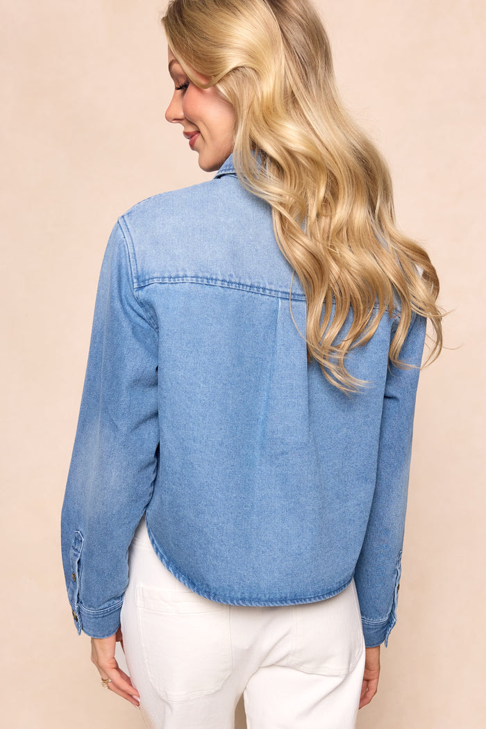 A woman with long, wavy blonde hair is shown from the back, wearing Ivy City Co’s Caroline Top in Light Wash Denim and white pants, standing against a beige background.