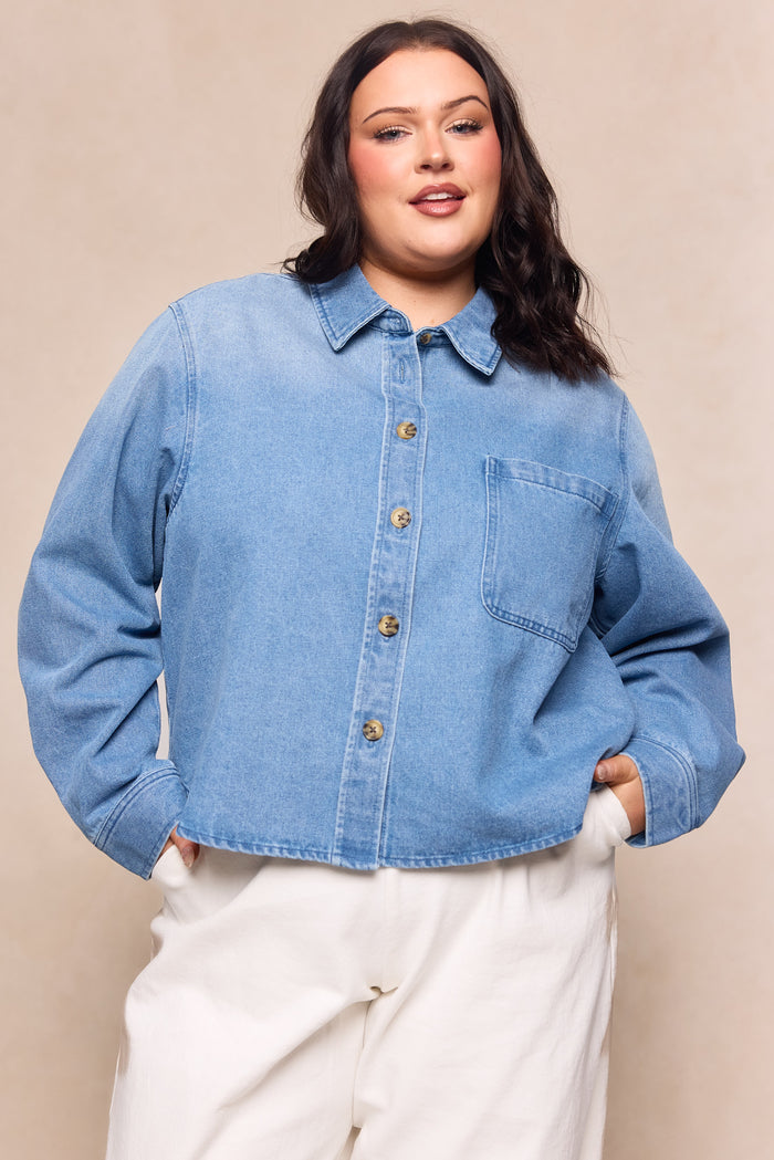 A woman with long brown hair smiles confidently with her hands in her pockets, wearing the Ivy City Co Caroline Top in Light Wash Denim and white pants, posed against a neutral background.