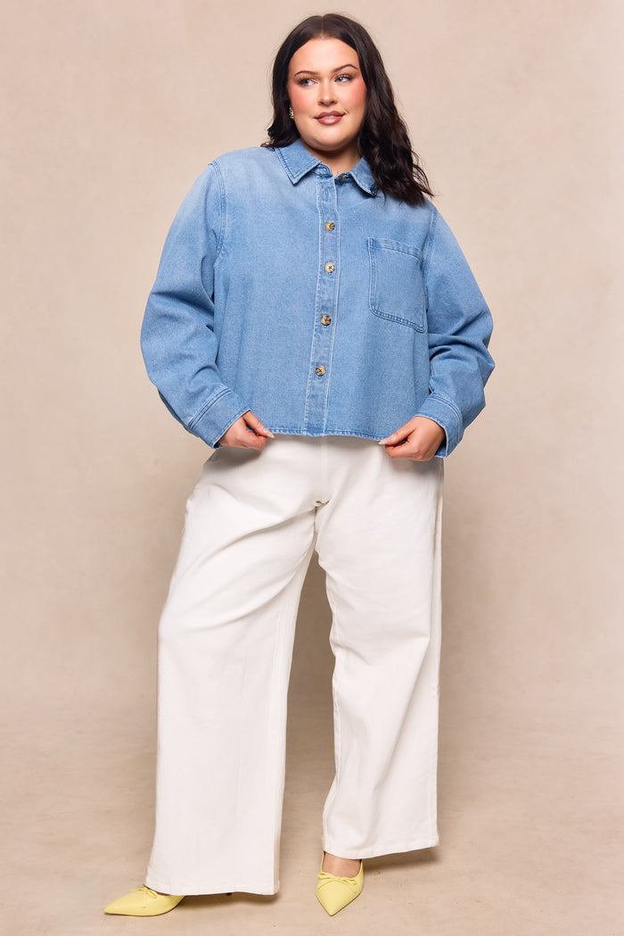 A woman with medium-length dark hair poses confidently against a light beige background, wearing the Ivy City Co Caroline Top in Light Wash Denim, paired with white wide-leg pants and yellow pointed-toe shoes.