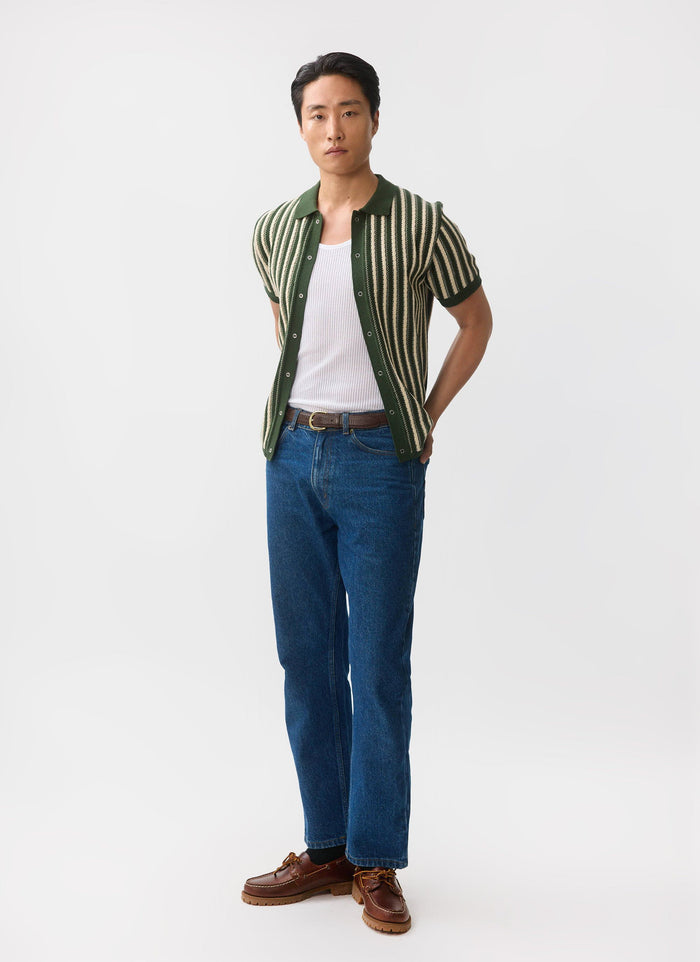 A man models the Percival Collins Micro Cable Knitted Shirt in green, layered over a white vest and styled with blue jeans.