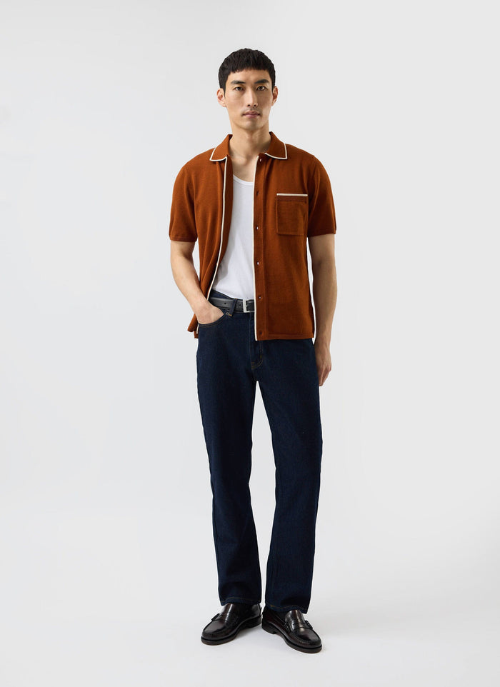 A model wears the Percival Contrast Trim Shirt in rust, a wool-blend knit styled open over a white vest and dark jeans.