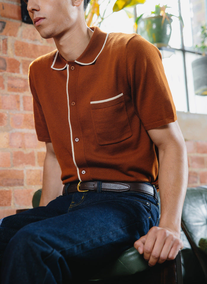 A model wears the Percival Contrast Trim Shirt in rust, a wool-blend knit with white piping, paired with dark jeans.
