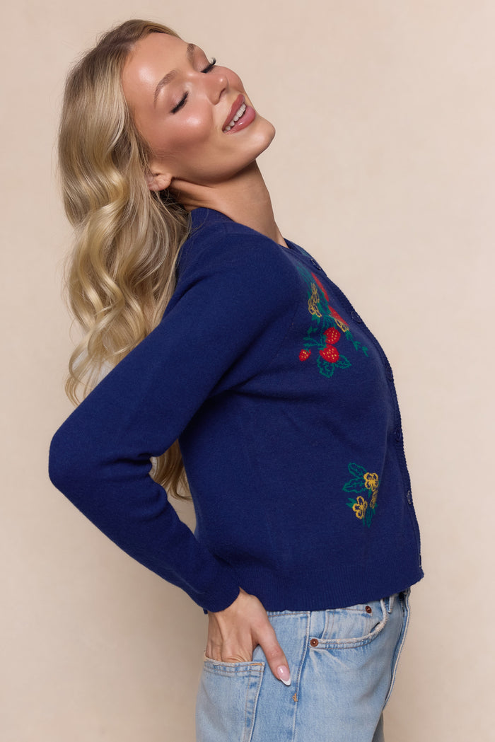 A woman with long, wavy blonde hair smiles with eyes closed, wearing the Ivy City Co Delphi Sweater in Strawberry Print and light blue jeans, standing against a beige background.