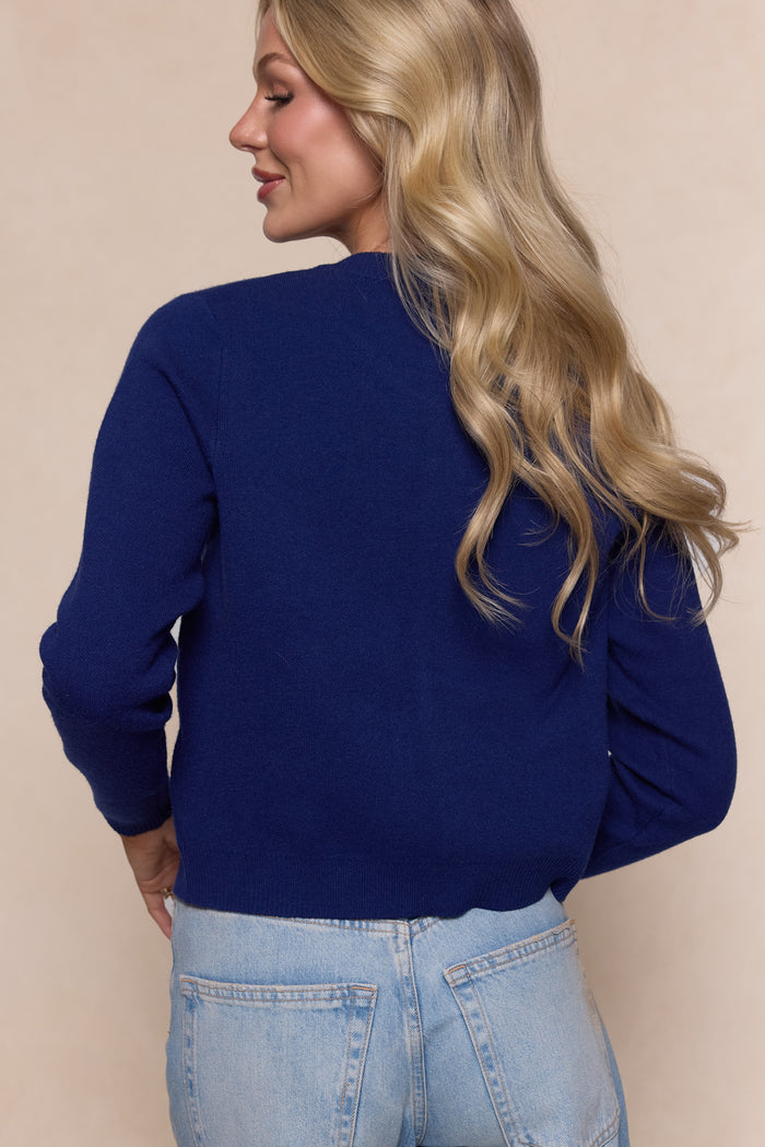 A woman with long, wavy blonde hair stands with her back to the camera, wearing Ivy City Co’s Delphi Sweater in Strawberry Print and light blue jeans against a neutral beige background.