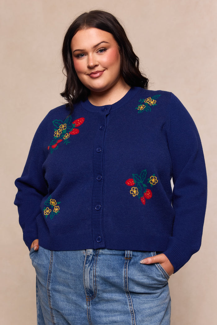 A woman with dark hair stands with her hands in her pockets, softly smiling at the camera while wearing the Ivy City Co Delphi Sweater in Strawberry Print, a maternity-friendly cardigan, paired with light blue jeans against a neutral background.