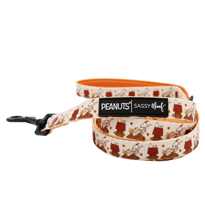 Cream-colored dog leash from the Sassy Woof Peanuts™ Harvest Three Piece Bundle, featuring a “Peanuts | Sassy Woof” label and adorable illustrations of Snoopy and Woodstock playing in autumn leaves. This leash has an orange interior, a durable black metal clasp, and pairs perfectly with the matching dog collar for the ultimate Puppy Growth Pawtection Plan.
