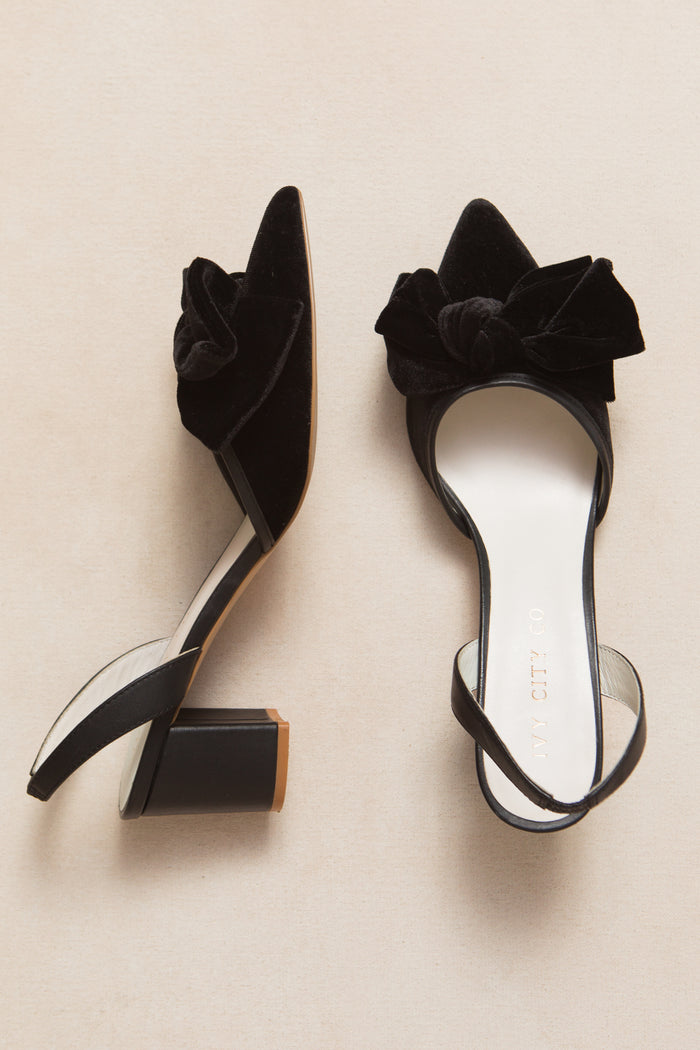The Eloise Bow Heel in Black by Ivy City Co features black velvet slingback heels with block heels and large matching bows on pointed toes, shown on a light beige surface—an elegant twist on the classic black bow heel.