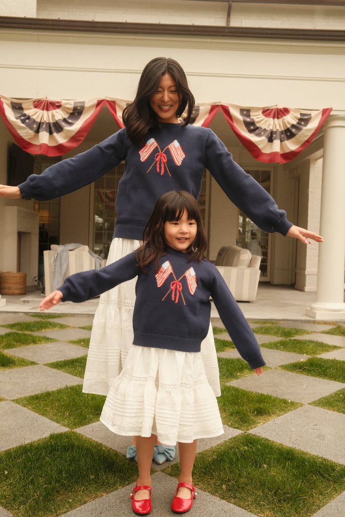 A woman and a young girl smile with arms outstretched, both wearing the Ivy City Co Freedom Sweater in Midnight with flag designs and white skirts, standing on a checkered patio decorated with patriotic bunting.