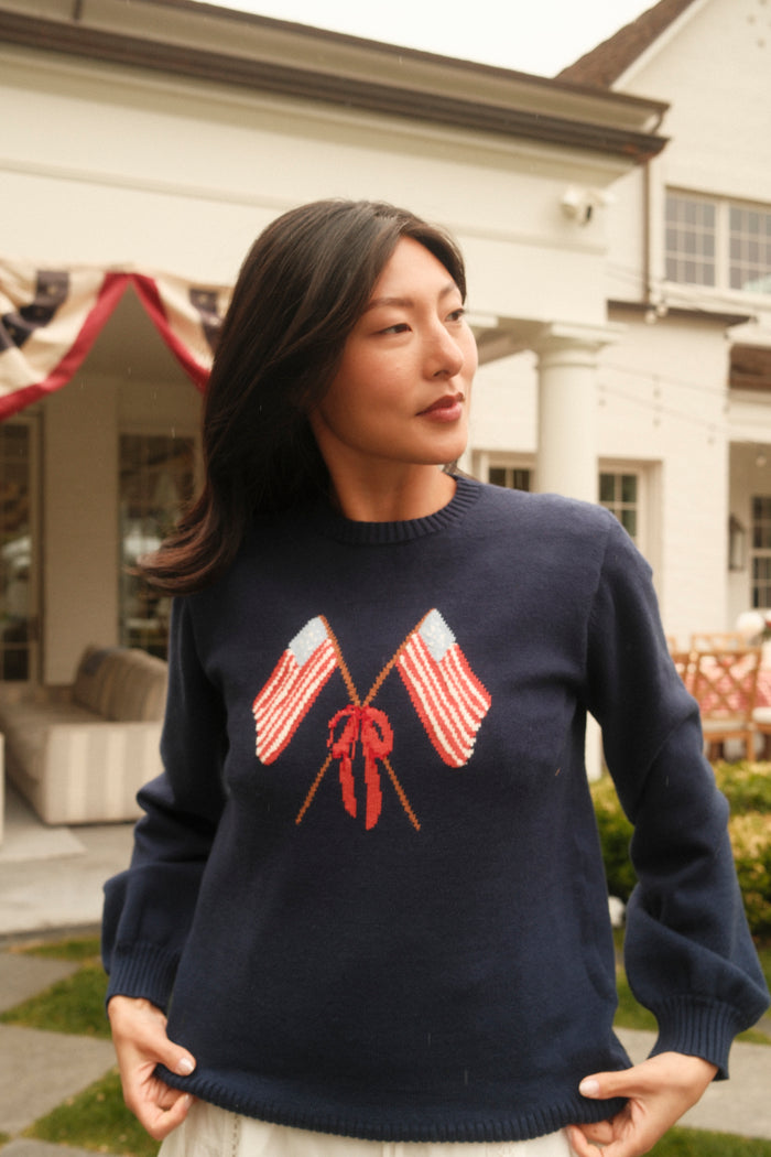 A woman stands outside a house wearing the Ivy City Co Freedom Sweater in Midnight, which features two crossed American flags and a red ribbon on the front. Patriotic bunting decorates the porch in the background.