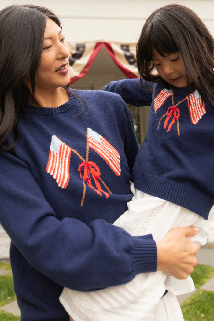 A woman and a young girl smile at each other outdoors, both in matching Ivy City Co Freedom Sweaters in Midnight with crossed American flags and red bows. Patriotic bunting decorates the background, adding to the festive scene.