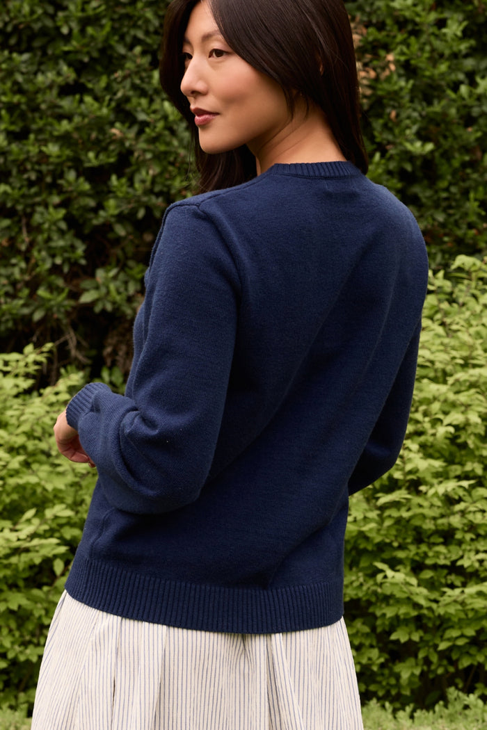 A woman with long dark hair stands outdoors among greenery, wearing the Ivy City Co Freedom Sweater in Midnight and a light-colored skirt, looking over her shoulder.