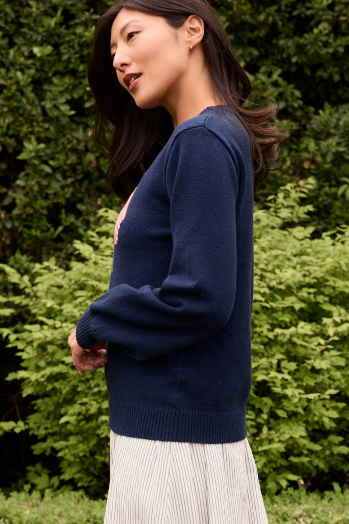 A woman with long dark hair wears the Ivy City Co Freedom Sweater in Midnight and a light pleated skirt, standing outdoors before lush green bushes, looking to the side with a slight smile.