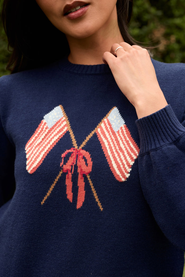 A woman wears the Ivy City Co Freedom Sweater in Midnight, featuring two crossed American flags above a red lobster. She touches her collar with one hand, with greenery visible in the background.