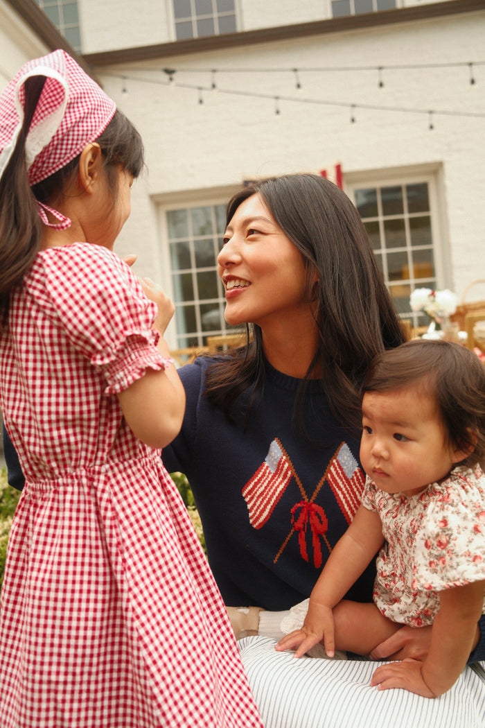 A woman wearing the Ivy City Co Freedom Sweater in Midnight smiles at a young girl in a red gingham dress and headscarf, holding a baby in a floral outfit on her lap outside near a building with large windows.