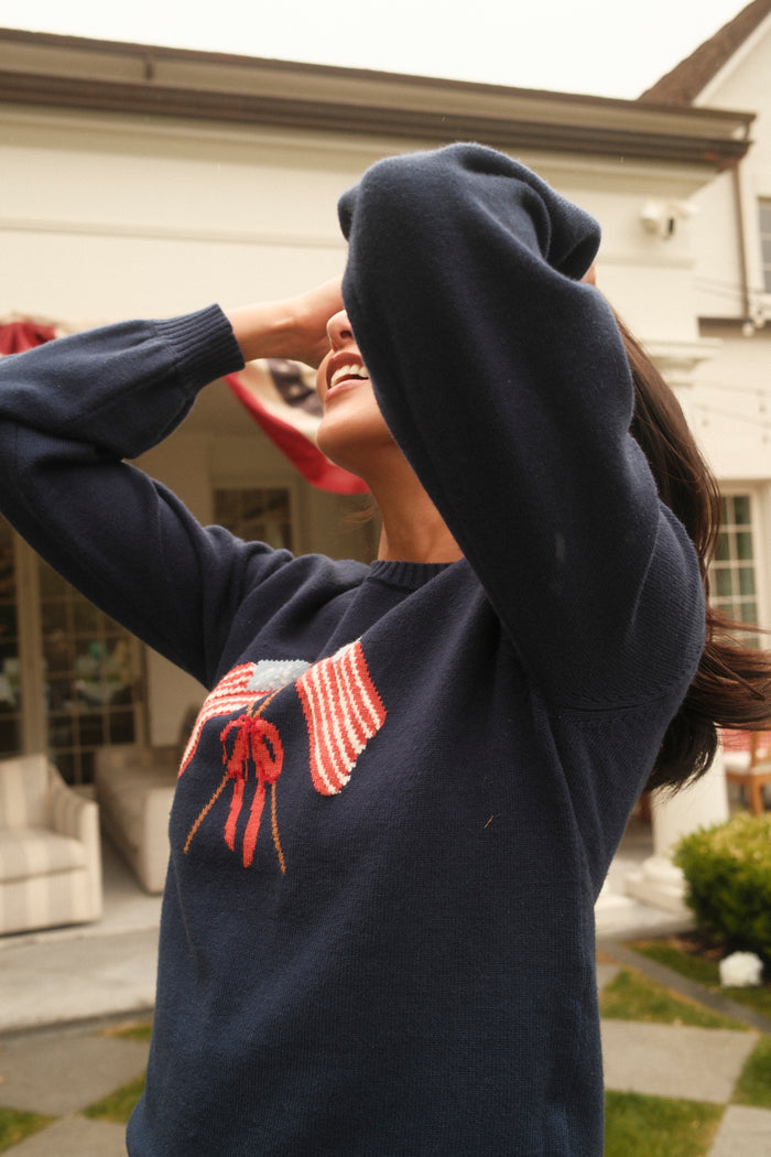 Outdoors, a smiling person covers their face with an arm and wears the Ivy City Co Freedom Sweater in Midnight, featuring two American flags and a red bow on the front. A house and greenery appear in the background.