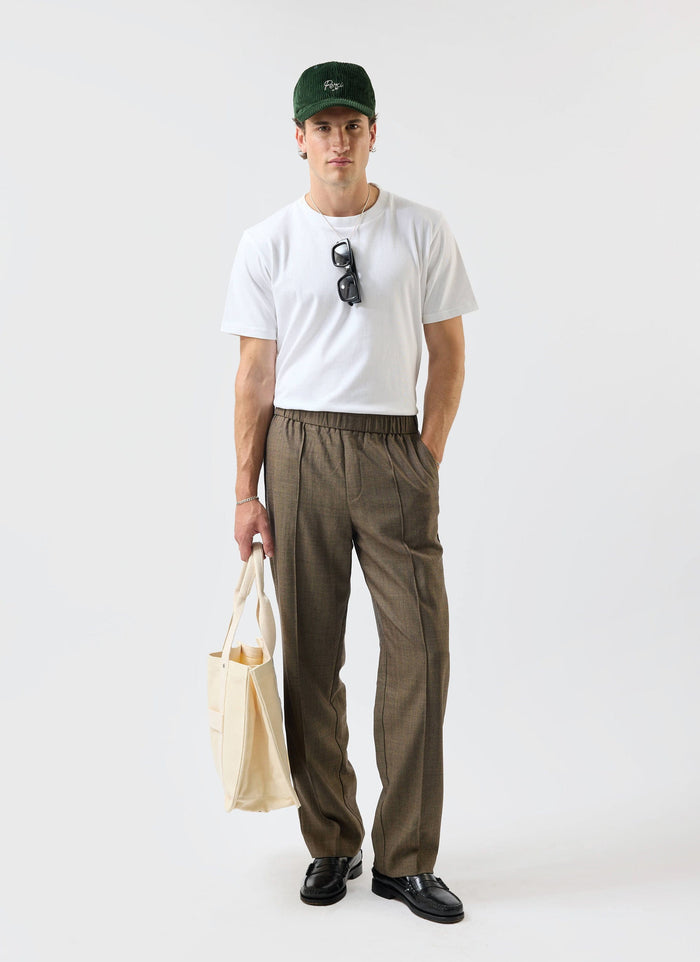 A man models the relaxed fit Percival Grounds Trousers in oak herringbone wool, paired with a white tee and loafers.