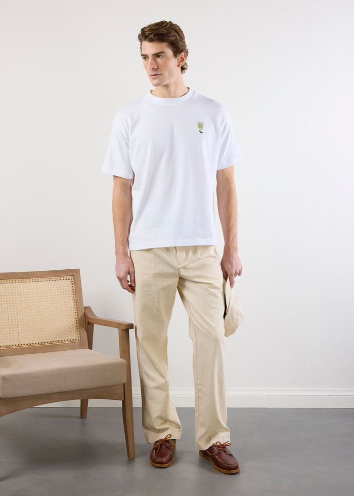 A man models the white Percival Iced Matcha t-shirt with beige trousers and brown boat shoes, standing beside a wooden chair.