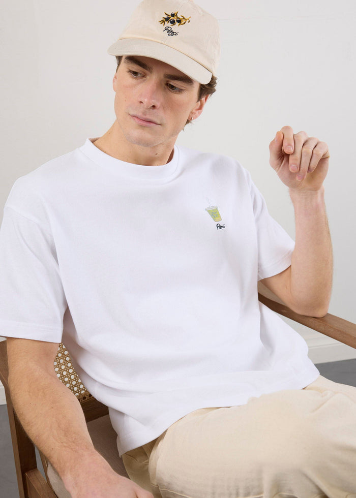 A model sits in a wooden chair wearing the white oversized Iced Matcha t-shirt with subtle chest embroidery by Percival.