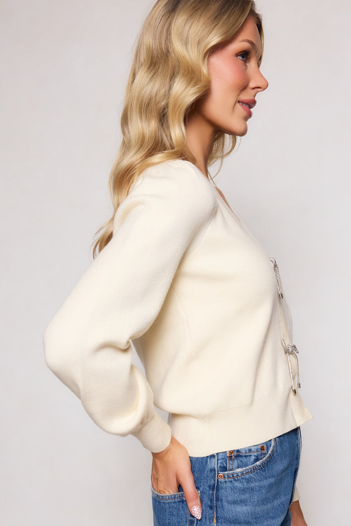 A woman with long wavy blonde hair wears the Illyria Sweater in Cream by Ivy City Co, styled with blue jeans. Posed in profile against a light background, she highlights the relaxed silhouette of this timeless knit piece.