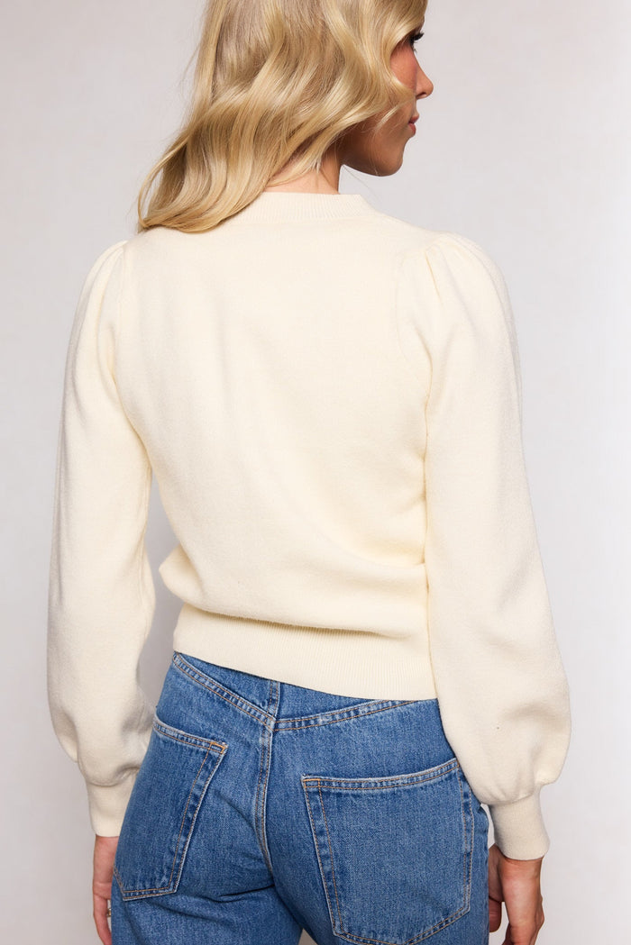 Wearing the Illyria Sweater in Cream by Ivy City Co, a woman with long blonde hair stands facing away from the camera. The sweater’s relaxed silhouette, featuring puff sleeves, pairs effortlessly with her blue jeans for easy everyday layering.