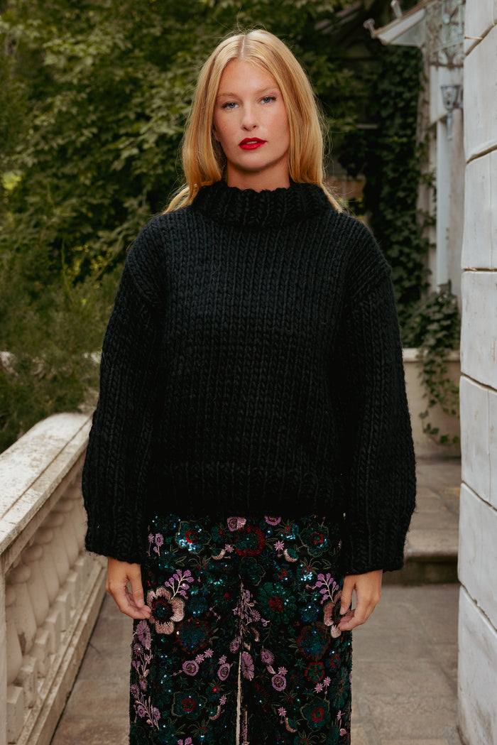 A person with long blonde hair wears the Ivy City Co Ira Sweater in Black and floral pants, standing outside near a white stone railing and lush green foliage.