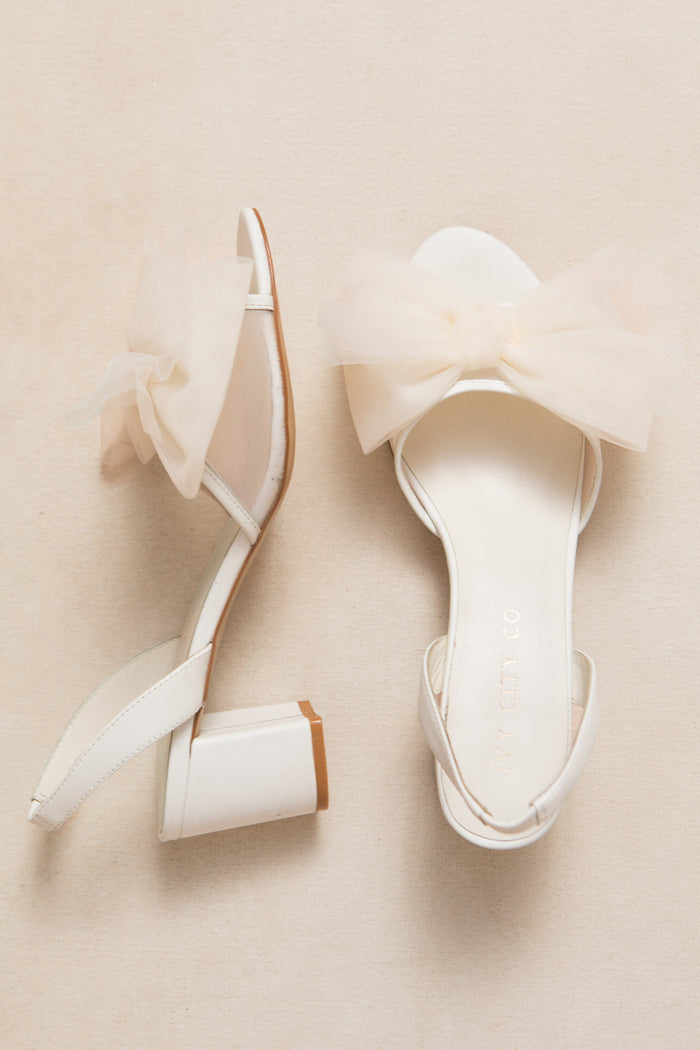 The Ivy Bow Heel in White by Ivy City Co features low heels, ankle straps, and sheer tulle bows on the front for a chic statement look.
