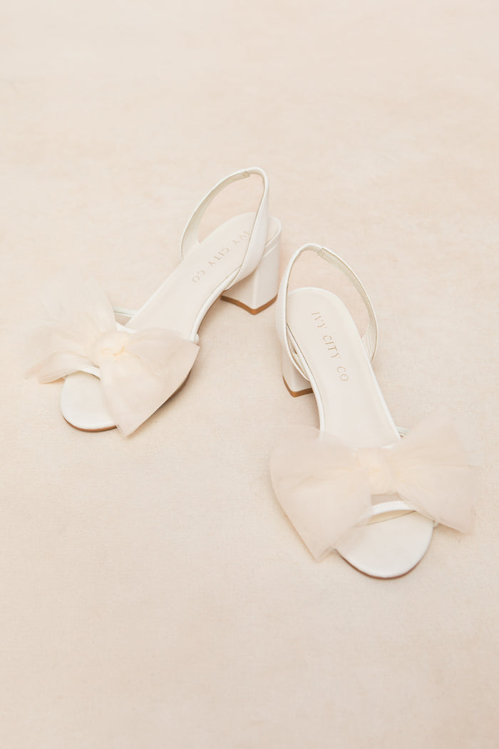 The Ivy Bow Heel in White by Ivy City Co features open toes, low heels, and large sheer tulle bows on the front straps, making a chic statement against a light beige background.