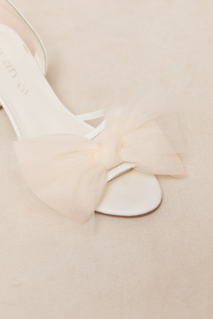 A close-up of the Ivy Bow Heel in White by Ivy City Co, a sandal with a sheer pale beige tulle bow on the front strap, shown resting on a soft, neutral surface.