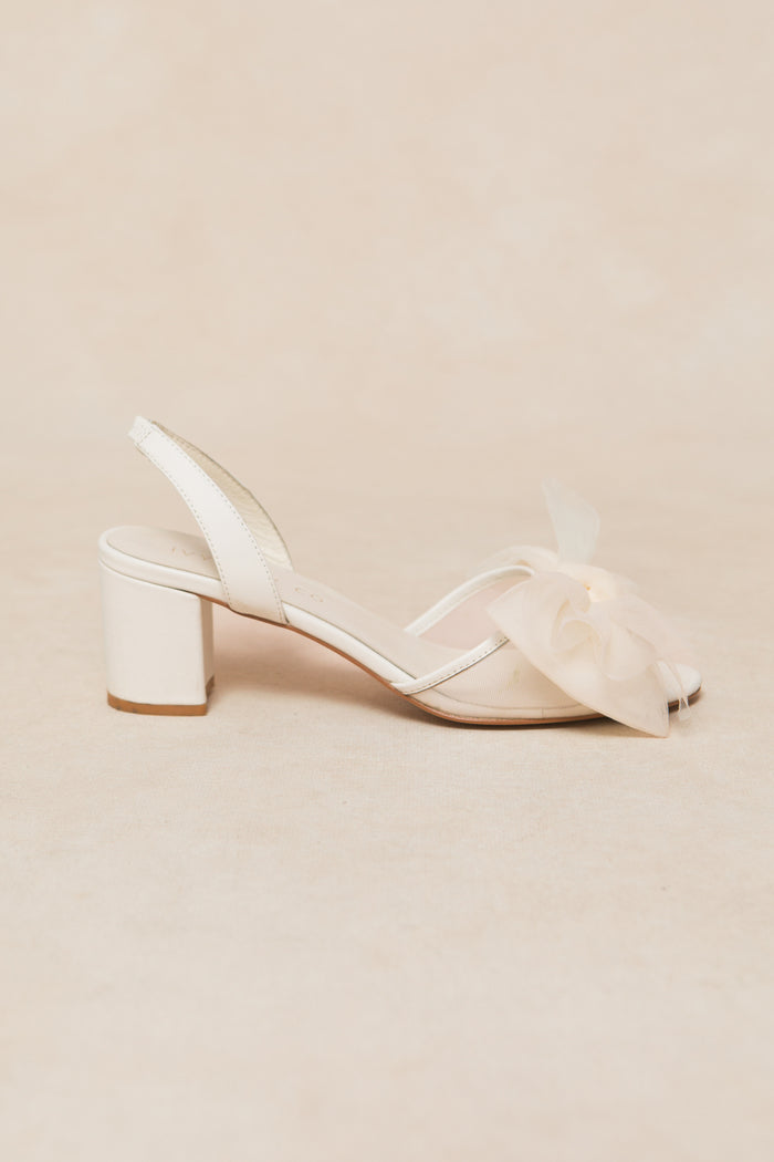 The Ivy Bow Heel in White by Ivy City Co features a single cream-colored tulle bow, an open toe, slingback strap, and a large sheer flower detail on the front, displayed against a neutral background.