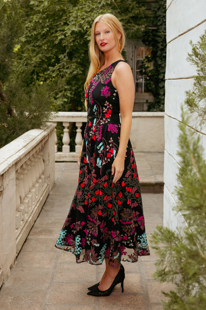 woman in multicolored one shoulder sleeve dress with embroidery standing on a patio with greenery behind