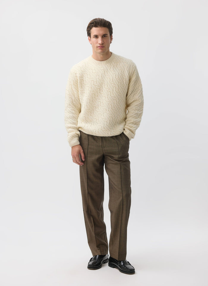 A model wears the Percival Melville Cable Knit Jumper in ecru, paired with smart brown trousers and black loafers.