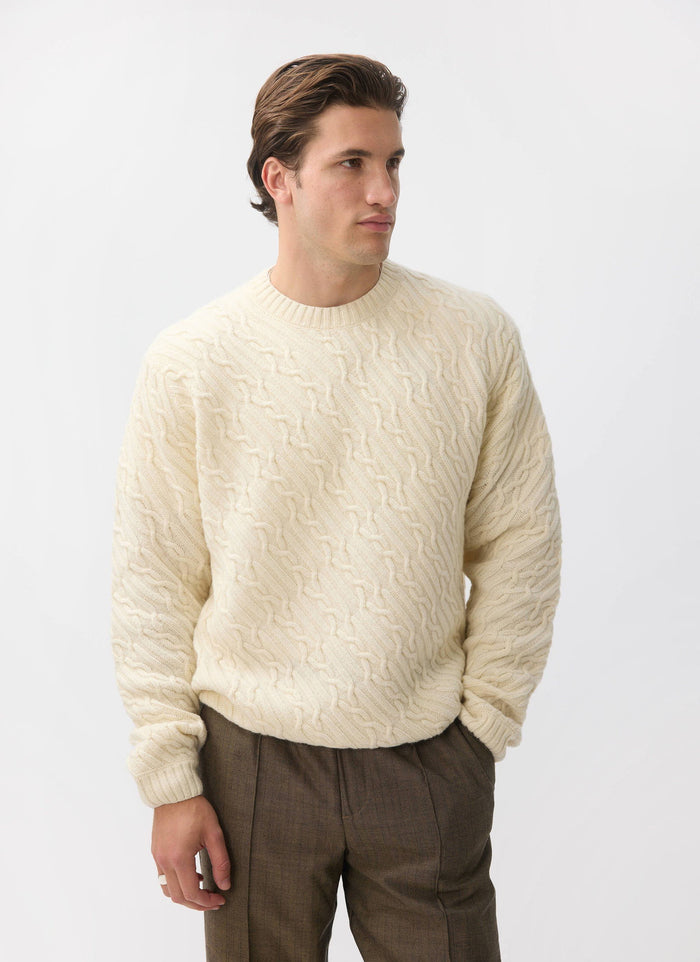 A model wears the Percival Melville Cable Knit Jumper in ecru, a wool-blend knit with a distinctive diagonal pattern.