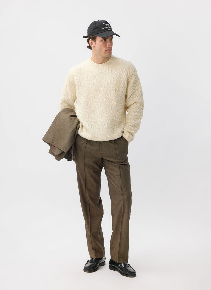A man models the Percival Melville Cable Knit Jumper in ecru, styled with brown trousers and a black cap.