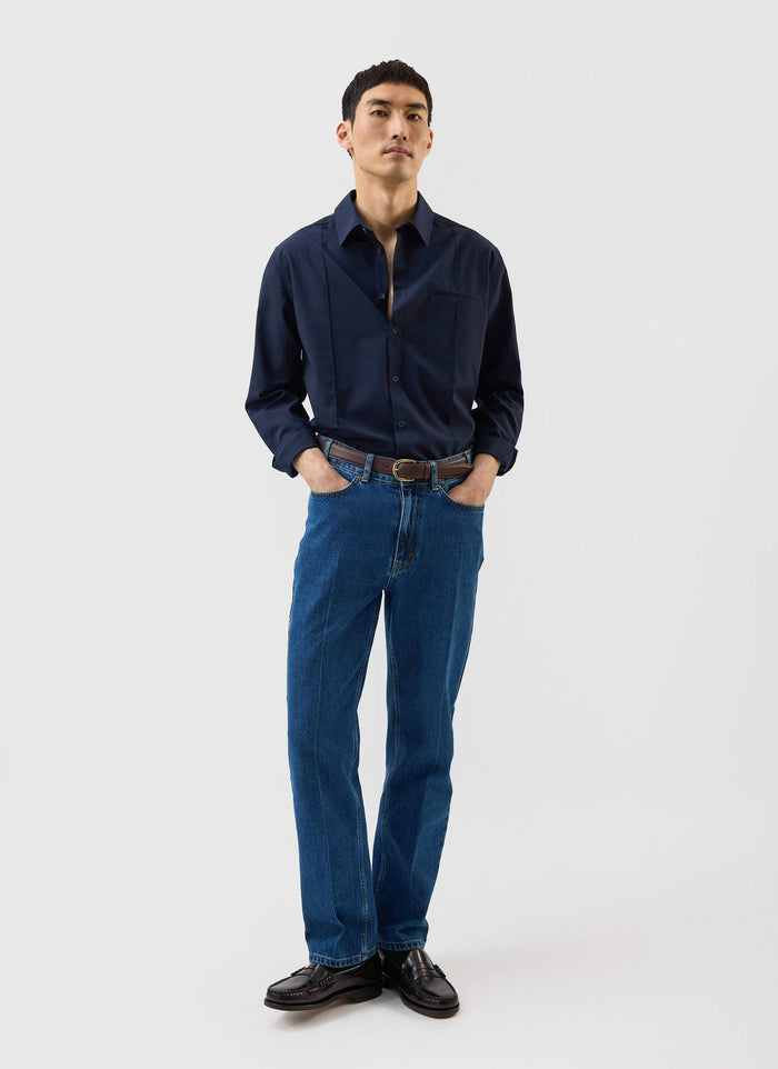 A model styles the Percival x Warren Pleated Shirt in navy blue with jeans and loafers against a white backdrop.