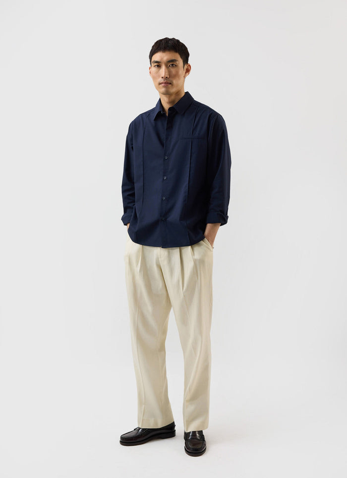 A man models the Percival x Warren navy blue pleated shirt, styled with cream trousers and black loafers.