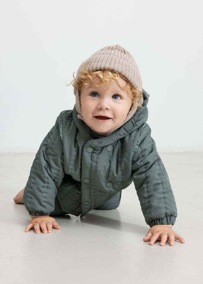 Baby wearing dusty green thermal jacket and hat, crawling on the floor