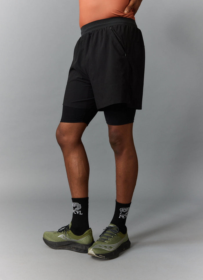 A side view of a model wearing the black PCVL 2 in 1 performance shorts from Percival with matching socks.