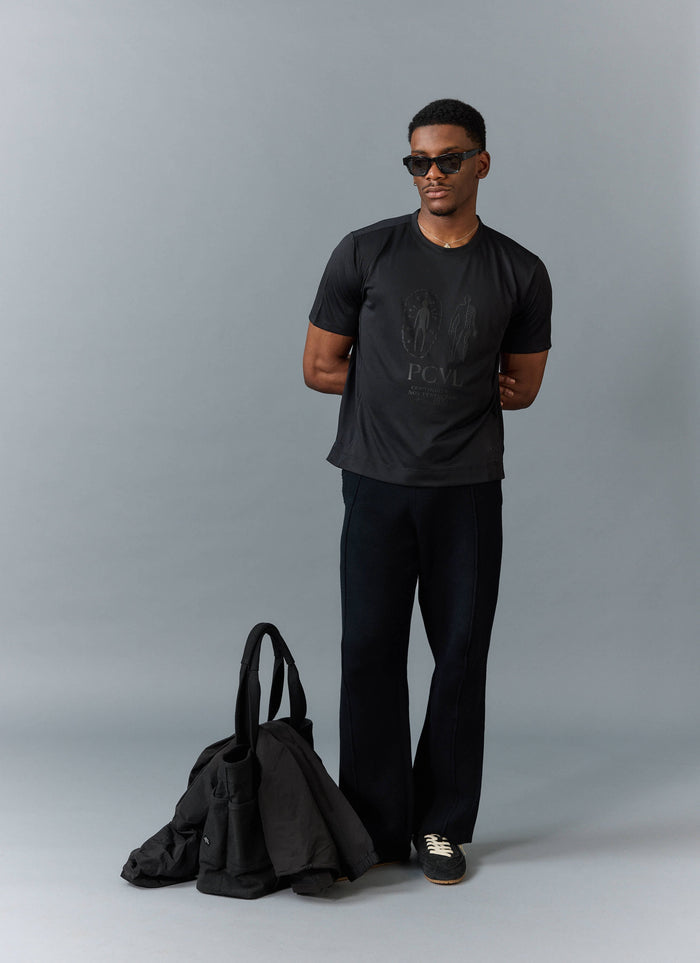 A model wears the black PCVL Printed Performance T-Shirt from Percival, styled with black trousers and sunglasses.